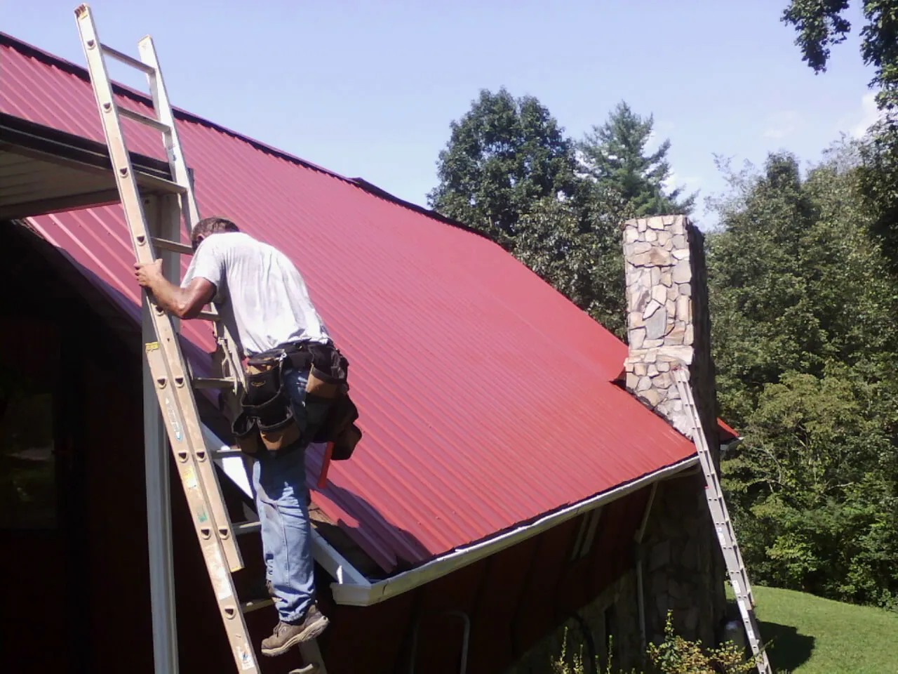 Roofer on a ladder beside a red metal roof and stone chimney — Soffit & Fascia Repair by Roofing Brothers in Spartanburg